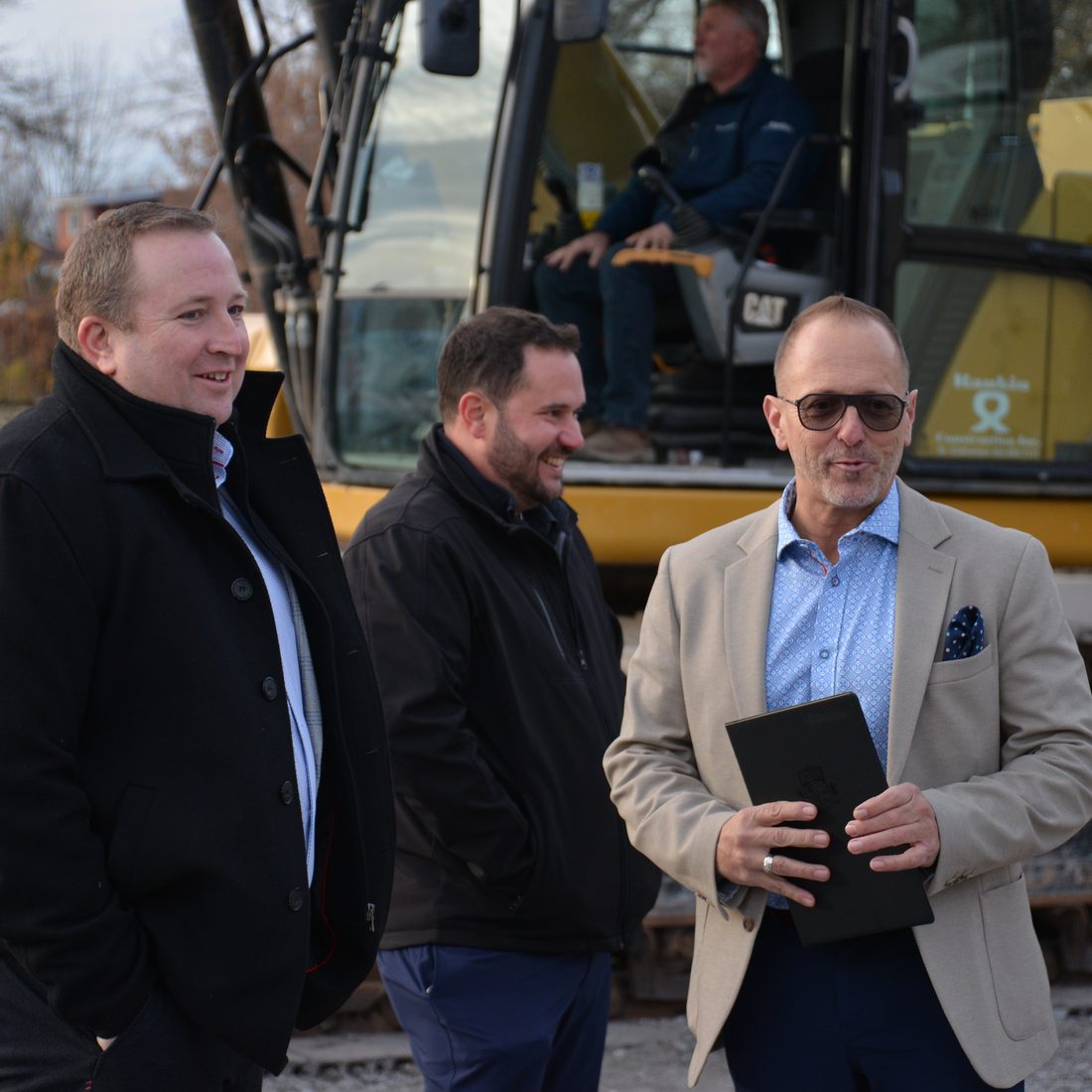 Three project partners in conversation beside a CAT excavator at the 4500 Park Street groundbreaking